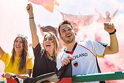 Das Bild zeigt zwei junge Frauen und einen Mann, die gemeinsam jubeln. Der Mann ist mit einem Deutschland-Fußballtrikot bekleidet. Die beiden Frauen haben sich die Wangen mit Deutschland Flaggen bemalt. Eine trägt ein gelbes T-Shirt, die andere hat ein schwar-rotes Shirt an. Die Frau links und der Mann rechts halten eine Deutschland Fahne. Verschwommen im Hintergrund sind weitere Personen erkennbar, die Fahnen schwenken und jubeln. 