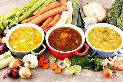 Drei Suppen stehen in Suppenschüsseln nebeneinander und darum ist Suppengemüse gelegt. 