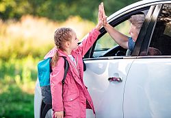Ein Mädchen steht neben der Fahrertür eines Autos und klatscht mit ihrem Vater durch das offene Fenster ab. 