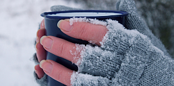 Hände mit grauen fingerlosen Handschuhen, auf die Schnee gefallen ist, halten eine blaue Blechtasse in der Hand. 