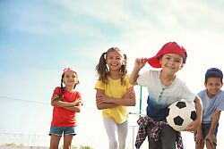 Fünf Kinder stehen auf einem Parkplatz. Einer trägt einen Fußball, die anderen sehen selbstbewusst in die Kamera. 