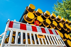 Nahaufnahme von gestapelten schwarzen Rohren mit gelben Endkappen hinter einer weißen Absperrung mit roten Streifen, im Hintergrund grüne Baumkronen.