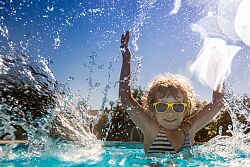 ein Kind im Freibad, mit gelber Sonnenbrille spritzt Wasser nach oben