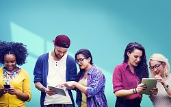 Zu sehen sind fünf junge Menschen, die jeweils ein Smartphone oder Tablet in den Händen halten und lachend darauf schauen. Die Gruppe besteht aus vier Frauen und einem jungen Mann. Alle tragen bunte Kelidung und wirken fröhlich unterhalten.