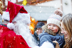 Die Stadt Lahr bietet originelle Weihnachtsgeschenke. &#13;&#10;&#13;&#10;Auf dem Bild steht der Weihnachtsmann in rotem Kostüm und weißem Bart in der Innenstadt und hält einem Kind seinen Jutesack hin. Das Kind ist auf dem Arm der Mutter und schaut den Weihnachtsmann mit großen Augen an. Vom Himmel fallen dicke Schneeflocken. 