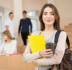 Stadt Lahr. Ein Mädchen steht in einem Hörsaal. Sie hat einen braunen Kaffeebecher in der Hand und vorne unter den Arm eine gelbe Mappe gepresst. Sie hat lange braune Haare und lächelt in die Kamera. Im Hintergrund sitzt ein weiteres Mädchen und zwei Jungs stehen. 