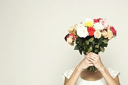 Zu sehen ist eine Frau in einem kurzen weißen Kleid, die einen bunten Blumenstrauß vor ihr Gesicht hält.