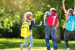 Drei Schulkinder sind auf einer Wiese, tragen Rucksäcke und springen jubelnd in die Luft. 