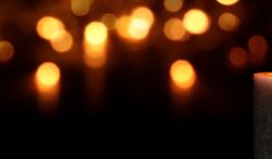 Zu sehen sind drei helle brennende Kerzen rechts im Vordergrund. Der Rest des Bildes ist dunkel und im Hintergrund erhellt von mehreren Lichtpunkten.