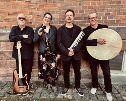 Auf dem Bild ist eine 4 köpfige Band zu sehen. Es sind drei Männer und eine Frau. Sie sind schwarz gekleidet. Jeder hat ein Instrument in den Händen.