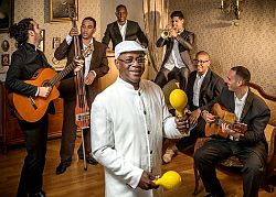Eine Gruppe kubanischer Musiker sitzen und stehen mit Ihren Instrumenten in einer Wohnzimmer-Kulisse.