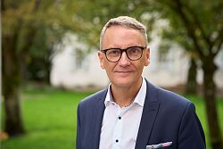 Mann mit grauen Haaren und Brille trägt weißen Hemdkragen und dunkelblauen Anzug vor unscharfem Hintergrund mit Bäumen und Gebäude.